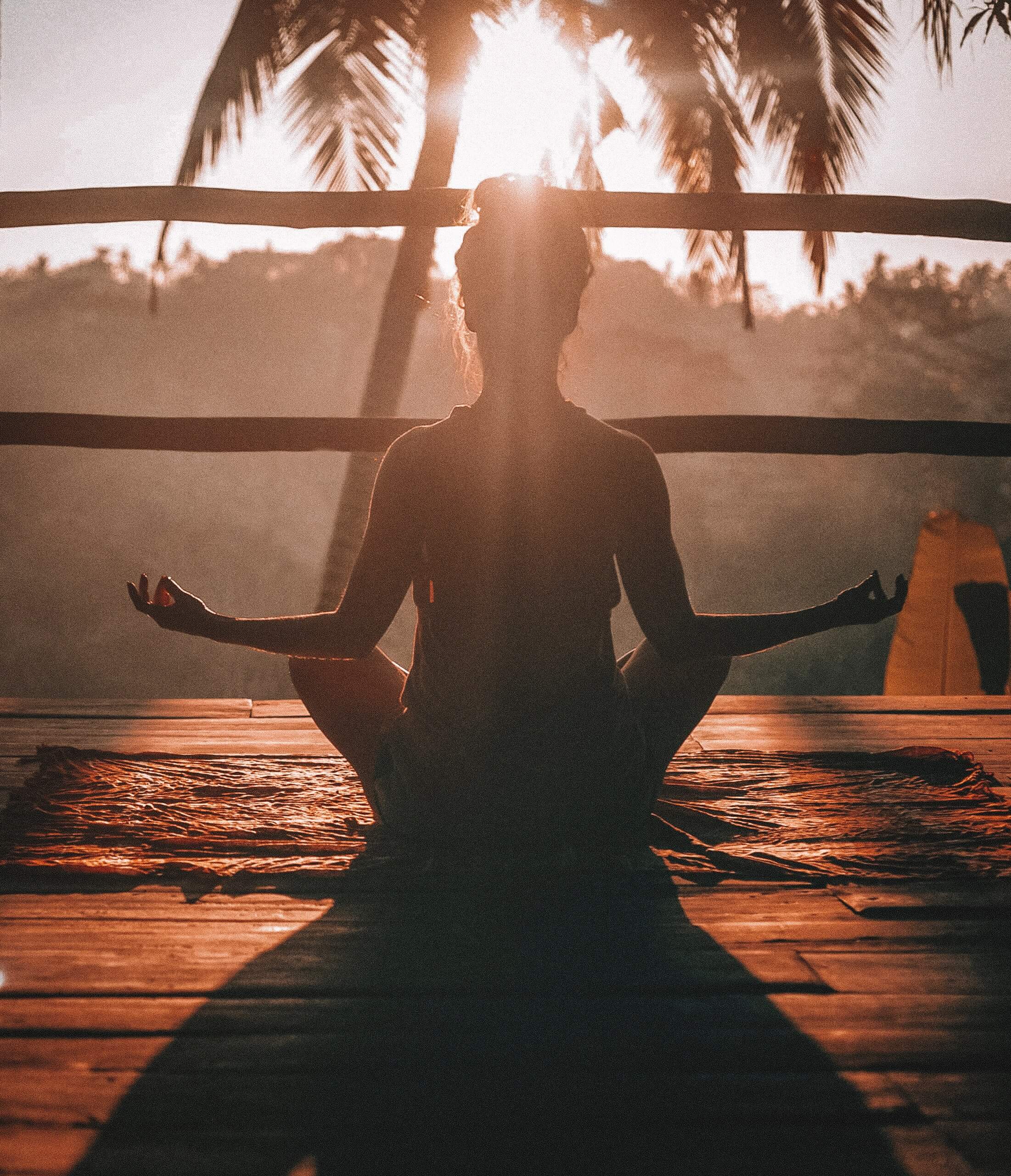 Sur une terrasse en bois, une jeune femme de dos en position assise de médiation lors du coucher de soleil devant un palmier